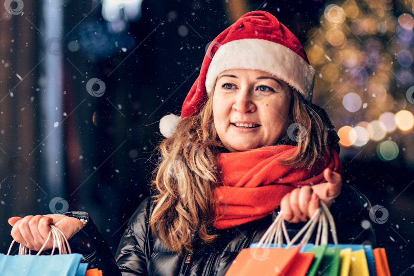 Скачать Портрет женщины в шляпе Санты с рождественскими покупками в разноцветных сумках на улице, украшенной рождественскими гирляндами и украшениями фотосток Ozero