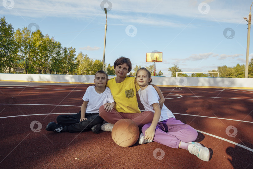 Скачать Объятия на открытом воздухе: Мать и дети-подростки нежно обнимаются на солнышке на баскетбольной площадке фотосток Ozero