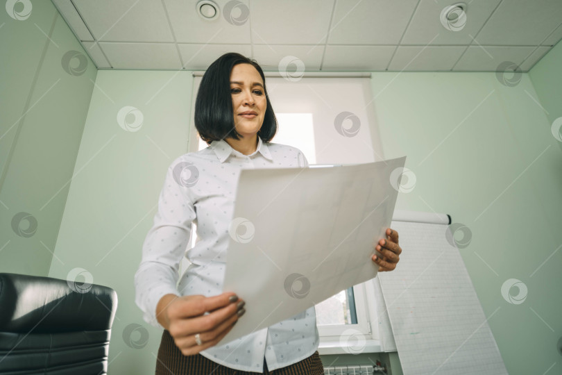 Скачать Портрет молодой деловой женщины, стоящей в офисе и смотрящей на графики, уверенно планирующей прибыль фотосток Ozero