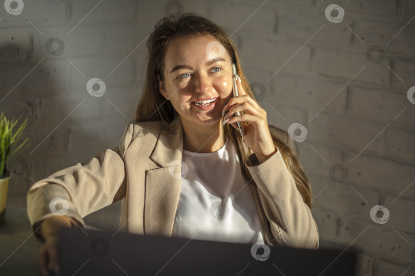 Скачать Улыбающаяся бизнесвумен разговаривает по телефону в офисе. Предприниматель малого бизнеса разговаривает по мобильному телефону и улыбается, работая за ноутбуком фотосток Ozero