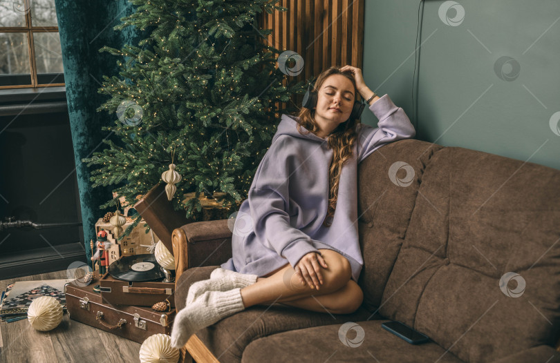 Скачать Молодая женщина в наушниках слушает музыку с виниловой пластинки, сидя на диване возле светящейся рождественской елки в День Рождества с закрытыми глазами. фотосток Ozero