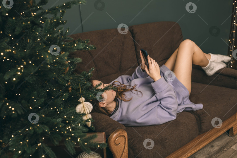 Скачать Неузнаваемая женщина лежит возле украшенной рождественской елки в гостиной на диване, улыбается и пользуется телефоном, набирая сообщение на смартфоне, празднуя новый год и Рождество. фотосток Ozero