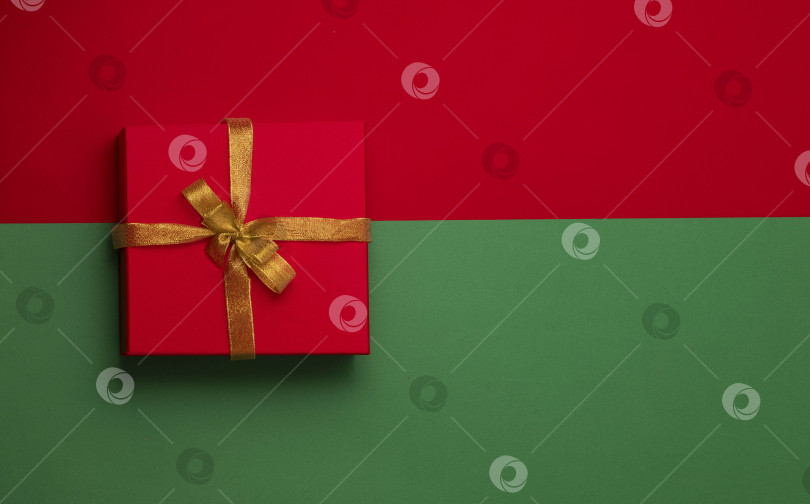 Скачать рождественская подарочная коробка красного цвета с золотым бантом на красно-зеленом фоне. Новогодняя концепция. Зимние праздники и подарки. Доставка-сюрприз фотосток Ozero