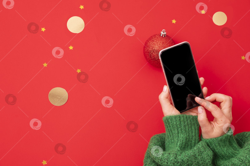 Скачать Женские руки держат мобильный телефон с конфетти из золотых звездочек и кружочков на фоне красной цветной бумаги в минималистском стиле, вид сверху фотосток Ozero