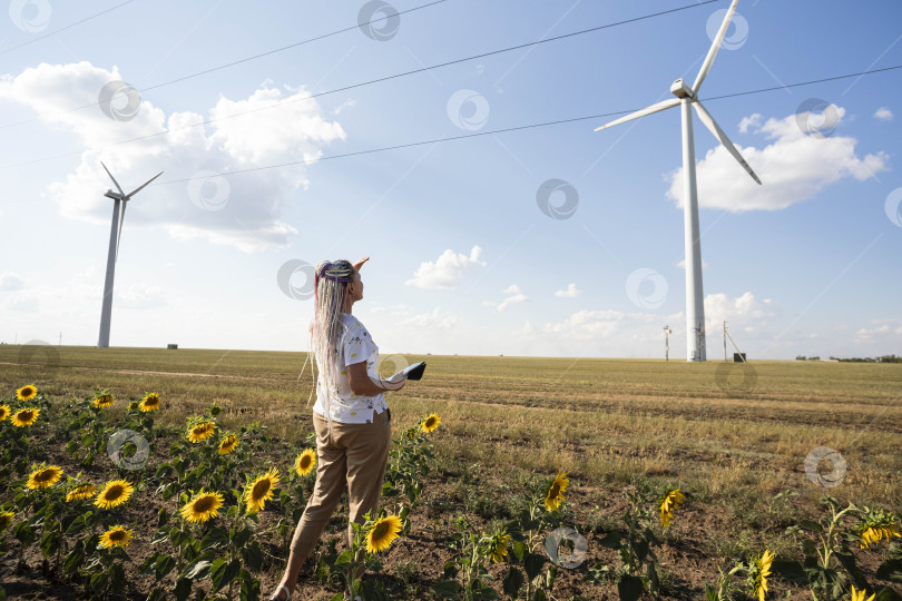 Скачать молодая женщина с планшетом в поле с подсолнухами изучает ветряные турбины для производства зеленой энергии, эко-энергия, ветряные турбины фотосток Ozero