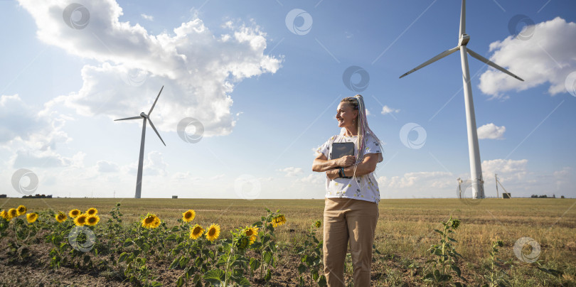 Скачать Молодая улыбающаяся женщина с планшетом в поле с подсолнухами, ветряные турбины для производства экологически чистой энергии, эко-энергия, ветряные турбины фотосток Ozero