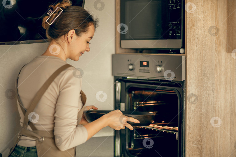 Скачать Молодая женщина в фартуке ставит блюдо в электрическую духовку для запекания на кухне фотосток Ozero