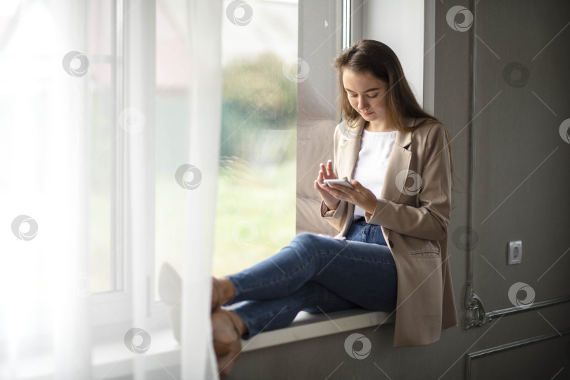 Скачать Баланс между работой и личной жизнью, белая женщина-менеджер сидит на подоконнике в холле со смартфоном фотосток Ozero