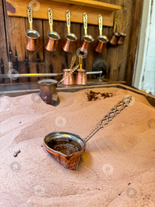 Скачать Медная джезва со свежим кофе по-турецки на песчаной плите, традиционные медные кофейники на деревянной подставке на заднем плане символизируют подлинность ближневосточных кофейных традиций. фотосток Ozero