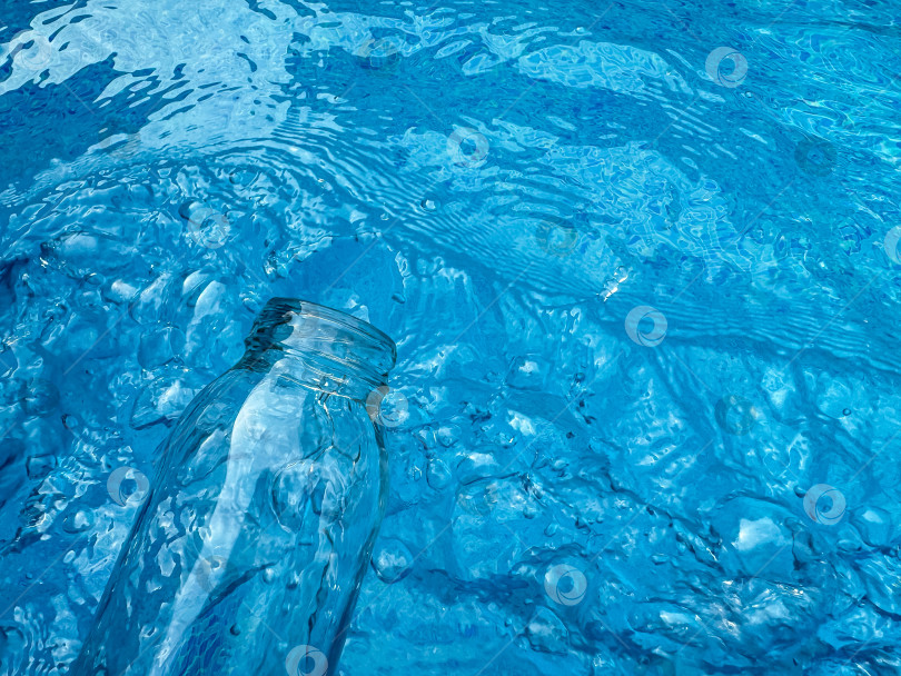 Скачать Бутылка из прозрачного стекла, частично погруженная в рябящую голубую воду с отражениями и преломлениями, вид сверху. Концепция чистой воды. фотосток Ozero
