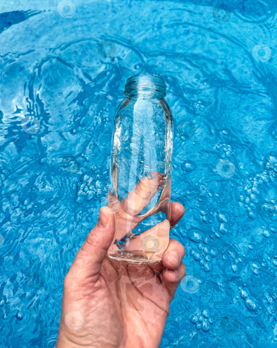 Скачать Рука, держащая прозрачную стеклянную бутылку, частично наполненную водой, на фоне сверкающей голубой воды. Концепция чистой воды. фотосток Ozero