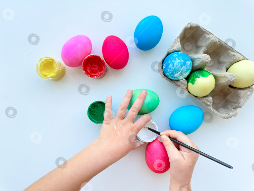 Скачать Детские ручки раскрашивают пасхальные яйца кисточкой в окружении разноцветных яиц и баночек с красками на белом столе - творческое праздничное занятие. фотосток Ozero