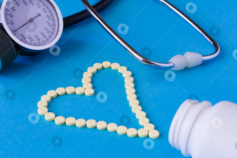 Скачать Желтые таблетки выложены в форме сердца, белая баночка и тонометр для измерения артериального давления со стетоскопом на синем фоне (концепция) фотосток Ozero