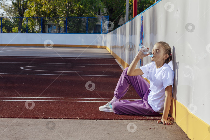 Скачать девушка пьет воду, сидя на резиновом покрытии баскетбольной площадки. Концепция спорта и активного образа жизни фотосток Ozero
