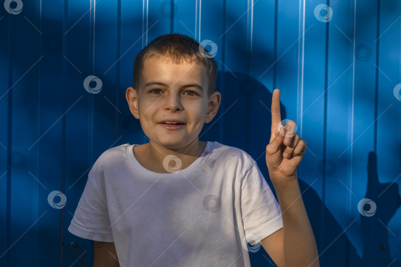 Скачать Маленький счастливый мальчик 10 лет в белой повседневной футболке, указывающий указательным пальцем в сторону на рабочем месте на простом синем фоне фотосток Ozero