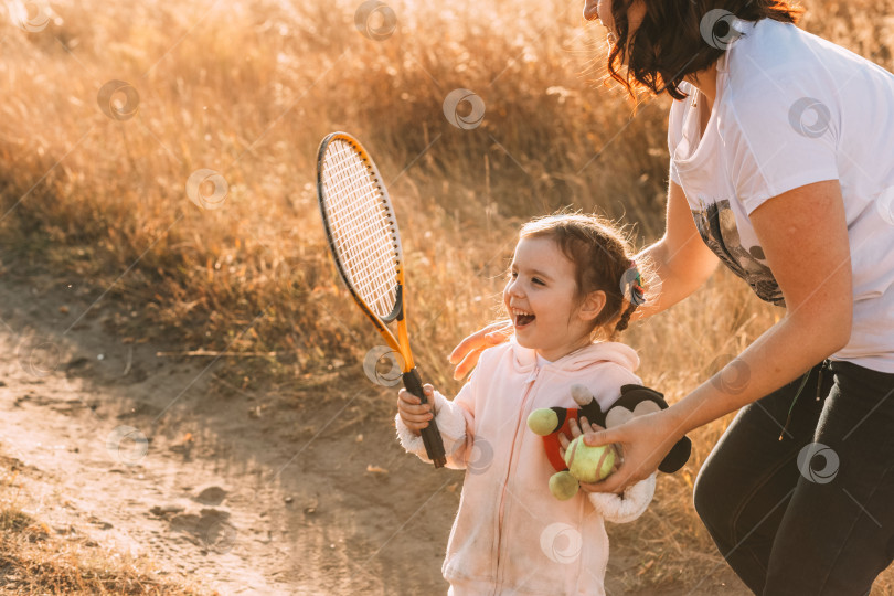 Скачать милая маленькая девочка наслаждается игрой с теннисной ракеткой в руке. Мама протягивает ей теннисный мяч фотосток Ozero