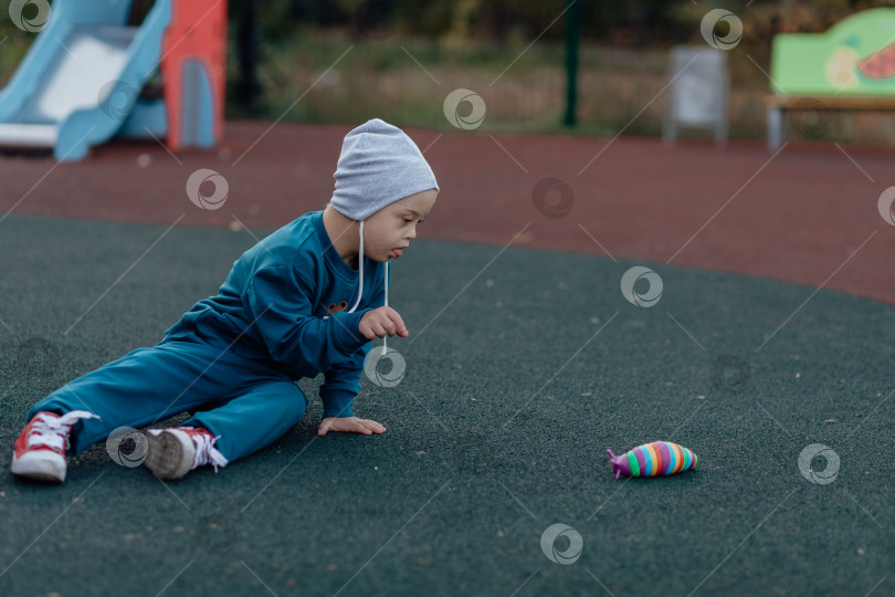 Скачать мальчик с синдромом Дауна сидит на резиновом покрытии на детской площадке и тянется за игрушкой фотосток Ozero