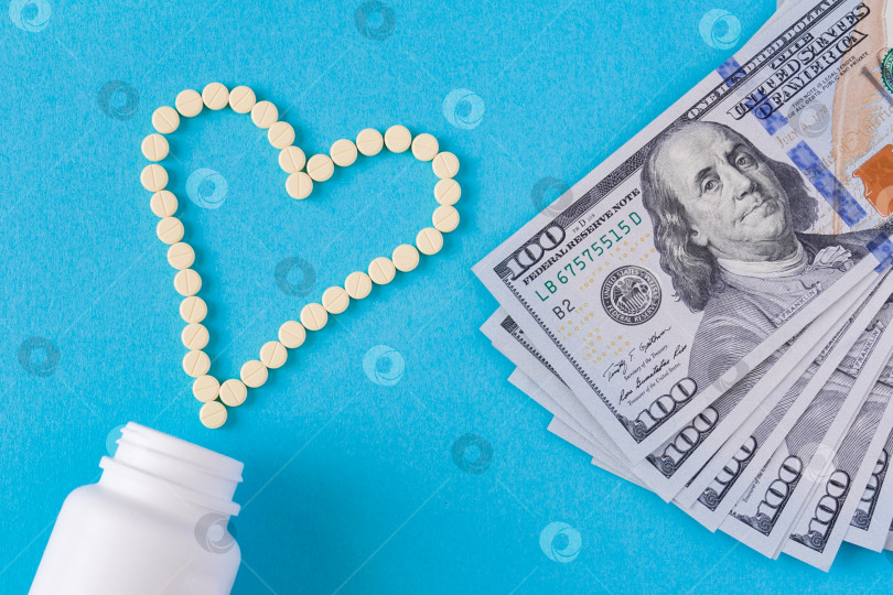 Скачать Желтые таблетки выложены в форме сердца. Белая банка и стодолларовые банкноты на синем фоне (концепция, плоское расположение) фотосток Ozero