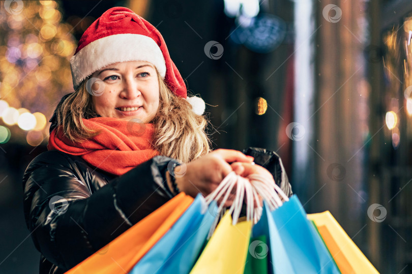 Скачать Портрет женщины в шляпе Санты с рождественскими покупками в разноцветных сумках на улице, украшенной рождественскими гирляндами и украшениями фотосток Ozero