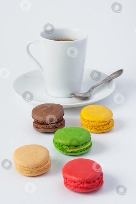 Скачать Французские разноцветные пирожные макароны на переднем плане и чашка кофе на блюдце с ложечкой в размытом виде на белом фоне фотосток Ozero