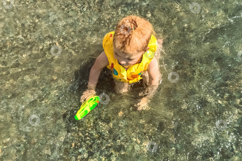 Скачать Маленькая девочка малыш сидит в жилете и с водяным пистолетом в воде на берегу моря фотосток Ozero