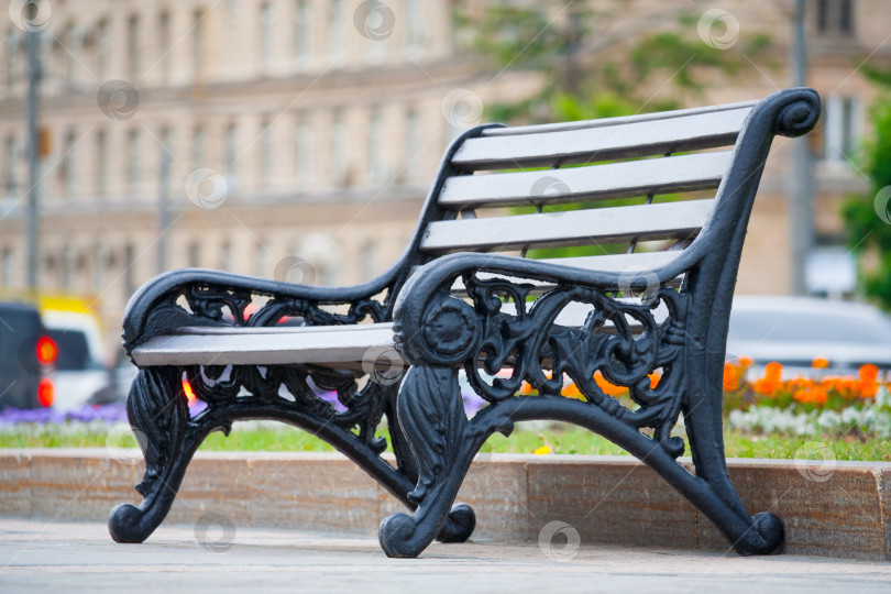 Скачать Крупный план серой деревянной скамейки с черными металлическими резными ножками в городском парке на фоне размытых зданий (малая глубина резкости) фотосток Ozero
