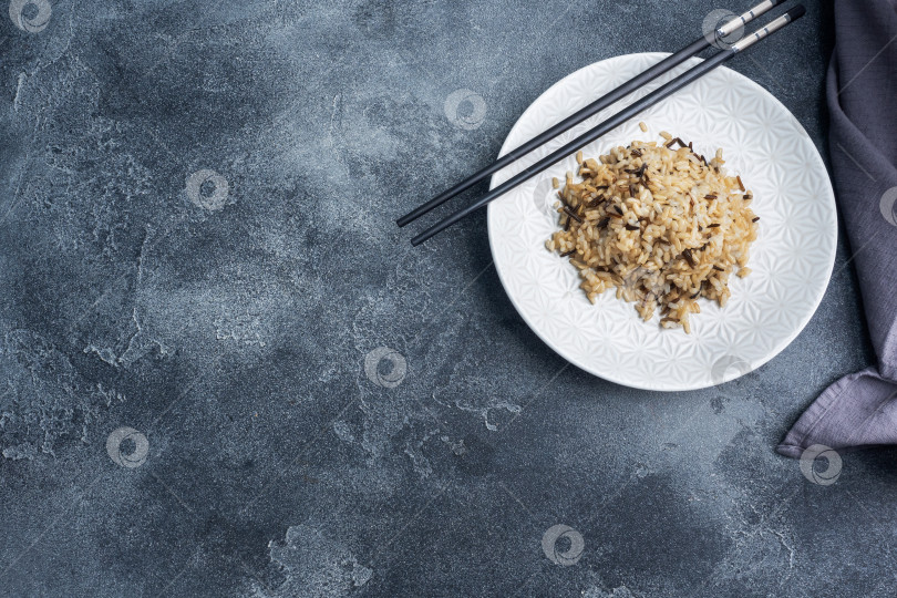 Скачать Обжаренный неочищенный рис на тарелке с палочками для еды. Темный бетонный фон. Скопируйте пробел. фотосток Ozero